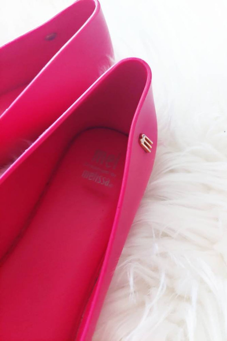 May include: A pair of bright pink ballet flats with a gold "M" logo on the inside of the shoe. The shoes are made of a rubber-like material and have a simple, sleek design.