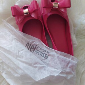 May include: A pair of pink ballet flats with a large bow on the toe. The shoes are made by Melissa and are packaged in a white tissue paper bag with the brand name printed on it.