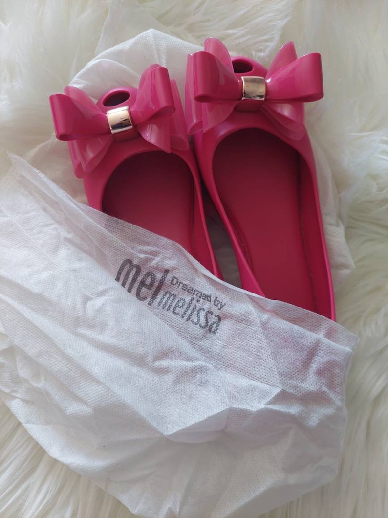 May include: A pair of pink ballet flats with a large bow on the toe. The shoes are made by Melissa and are packaged in a white tissue paper bag with the brand name printed on it.