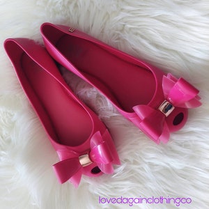 May include: A pair of pink ballet flats with a large bow on the toe. The shoes are made of a rubber-like material and have a gold accent on the bow.