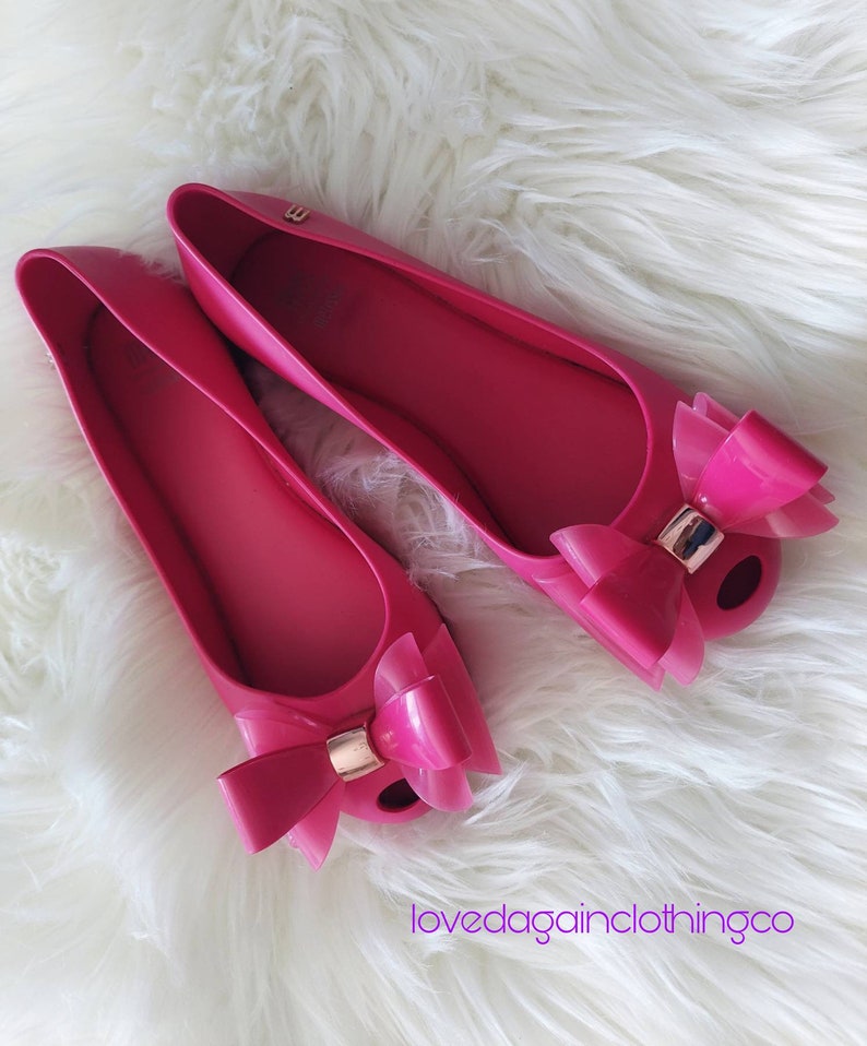 May include: A pair of pink ballet flats with a large bow on the toe. The shoes are made of a rubber-like material and have a gold accent on the bow.