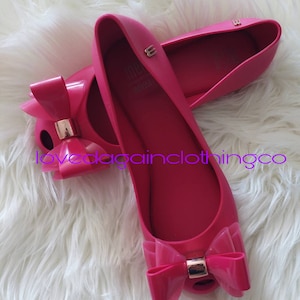 May include: A pair of pink ballet flats with a large bow on the toe. The shoes are made of a rubbery material and have a gold accent on the bow.