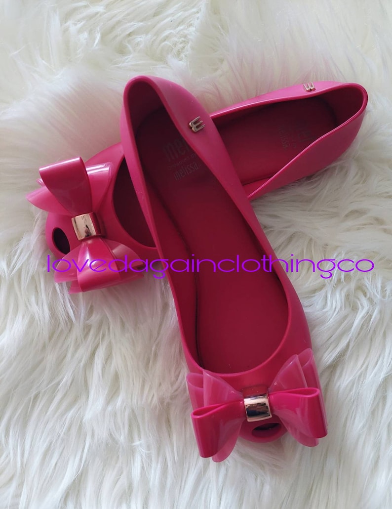 May include: A pair of pink ballet flats with a large bow on the toe. The shoes are made of a rubbery material and have a gold accent on the bow.