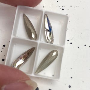 Op de afbeelding: Vier gefacetteerde, druppelvormige strass-steentjes in een witte, verdeelde tray. De strass-steentjes zijn een mix van zilver en grijs, met een reflecterende, metallic afwerking. De tray wordt door een hand vastgehouden en de achtergrond is wit met zwarte spikkels.