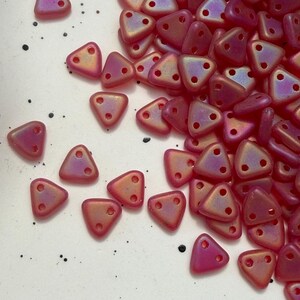 May include: A pile of iridescent triangular beads in pink, purple, and gold hues. Each bead features two holes for threading. The beads are scattered on a white surface with black speckles.