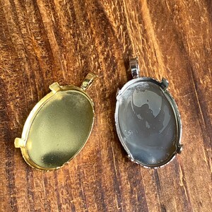 May include: Two oval-shaped metal pendant trays with a silver and gold finish. The trays have a loop at the top for attaching a chain or cord.