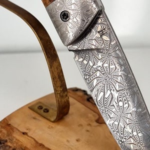 Może przedstawiać: Składany nóż ze stalową, wzorzystą klingą i drewnianym uchwytem. Uchwyt wykonany jest z drewna korzeniowego w naturalnym brązowym kolorze. Klinga ozdobiona jest spiralnym wzorem.