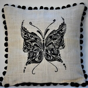 May include: Square decorative pillow with a natural linen-like fabric. The pillow features a large, intricate black butterfly design in the center. The edges are trimmed with black pom-poms, adding a decorative touch to the pillow.