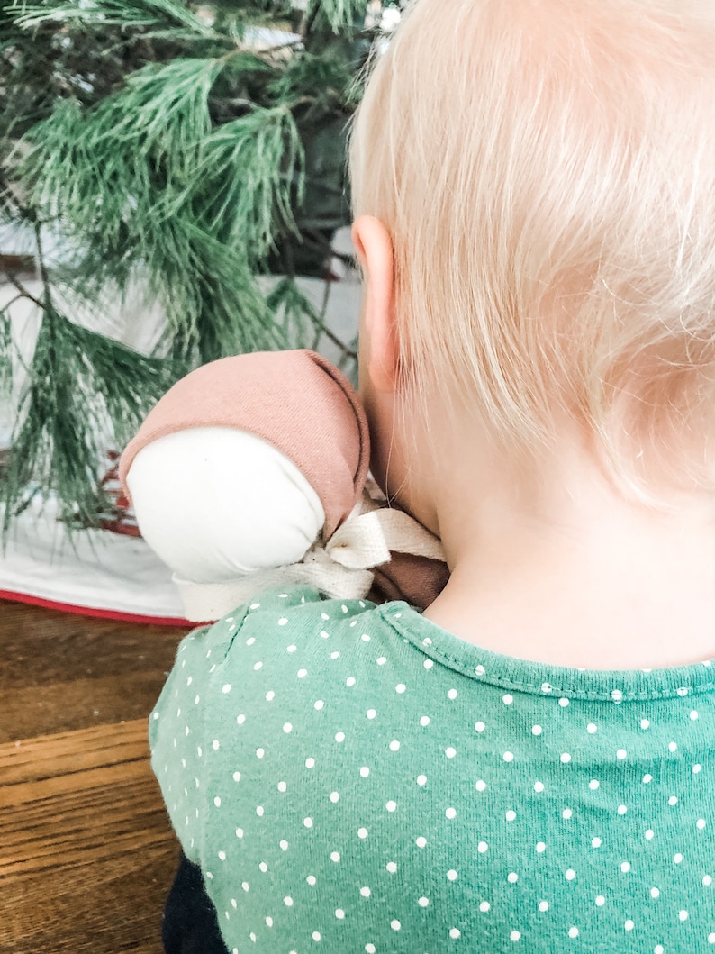 May include: A young child with blonde hair wearing a green shirt with white polka dots holds a pink and white fabric doll.
