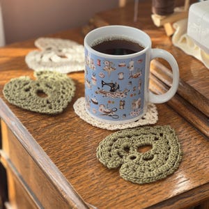 May include: A blue ceramic mug with a sewing themed design, filled with dark coffee, sitting on a wooden surface with three crocheted heart-shaped coasters. Two of the coasters are green and one is cream.