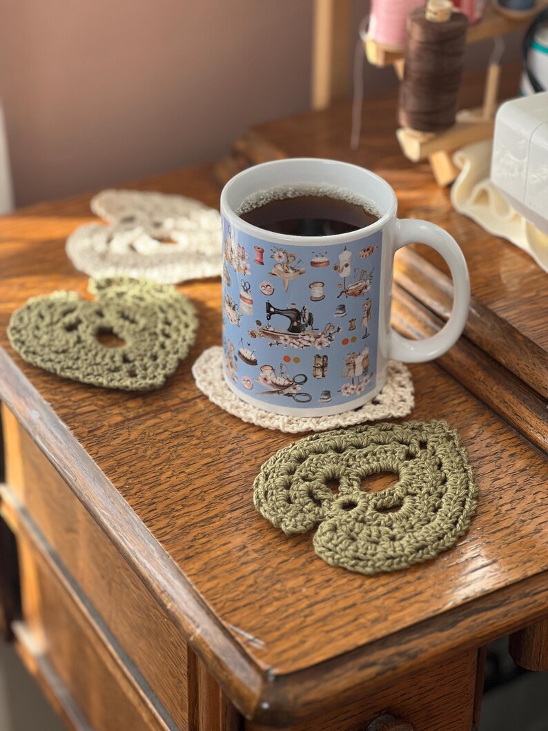 May include: A blue ceramic mug with a sewing themed design, filled with dark coffee, sitting on a wooden surface with three crocheted heart-shaped coasters. Two of the coasters are green and one is cream.