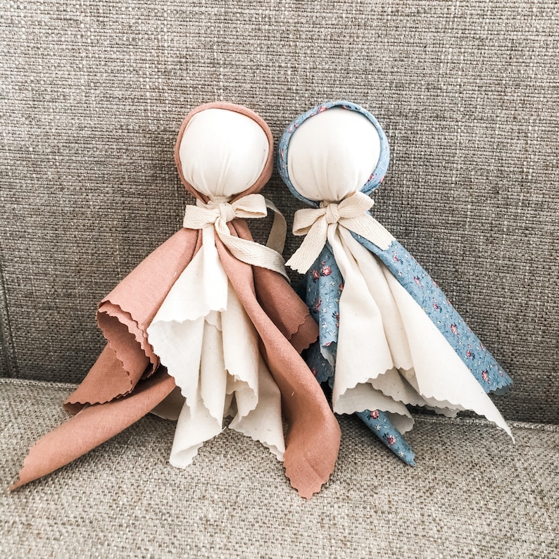 May include: Two handmade fabric dolls with white heads and fabric bodies. One doll has a brown fabric body with white fabric accents and the other doll has a blue fabric body with white fabric accents.