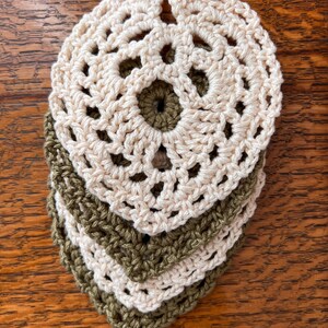 May include: Four crocheted coasters, two in a light beige color and two in a dark olive green color. The coasters are stacked on top of each other, with the beige coasters on top.