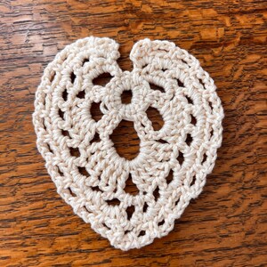 May include: A crocheted heart-shaped doily in a cream color. The doily is made with a delicate stitch pattern and has a scalloped edge.