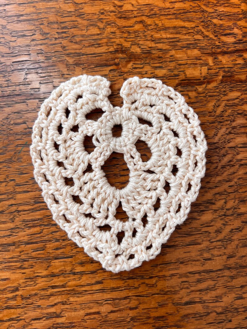 May include: A crocheted heart-shaped doily in a cream color. The doily is made with a delicate stitch pattern and has a scalloped edge.