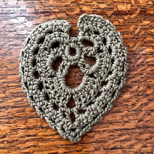 May include: A crocheted green leaf-shaped coaster with a detailed design. The coaster is made with a single crochet stitch and has a hole in the center.