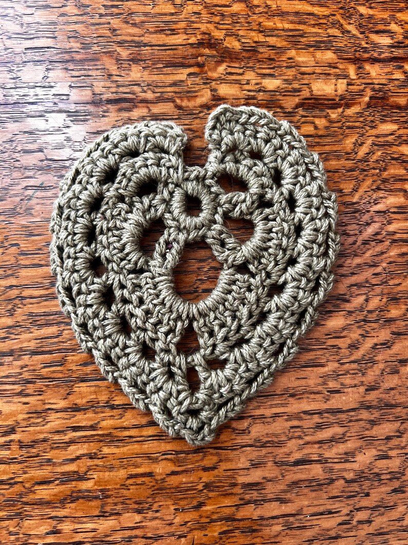 May include: A crocheted green leaf-shaped coaster with a detailed design. The coaster is made with a single crochet stitch and has a hole in the center.