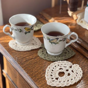 May include: Two white teacups with floral designs and brown tea inside, sitting on crocheted heart-shaped coasters on a wooden surface.