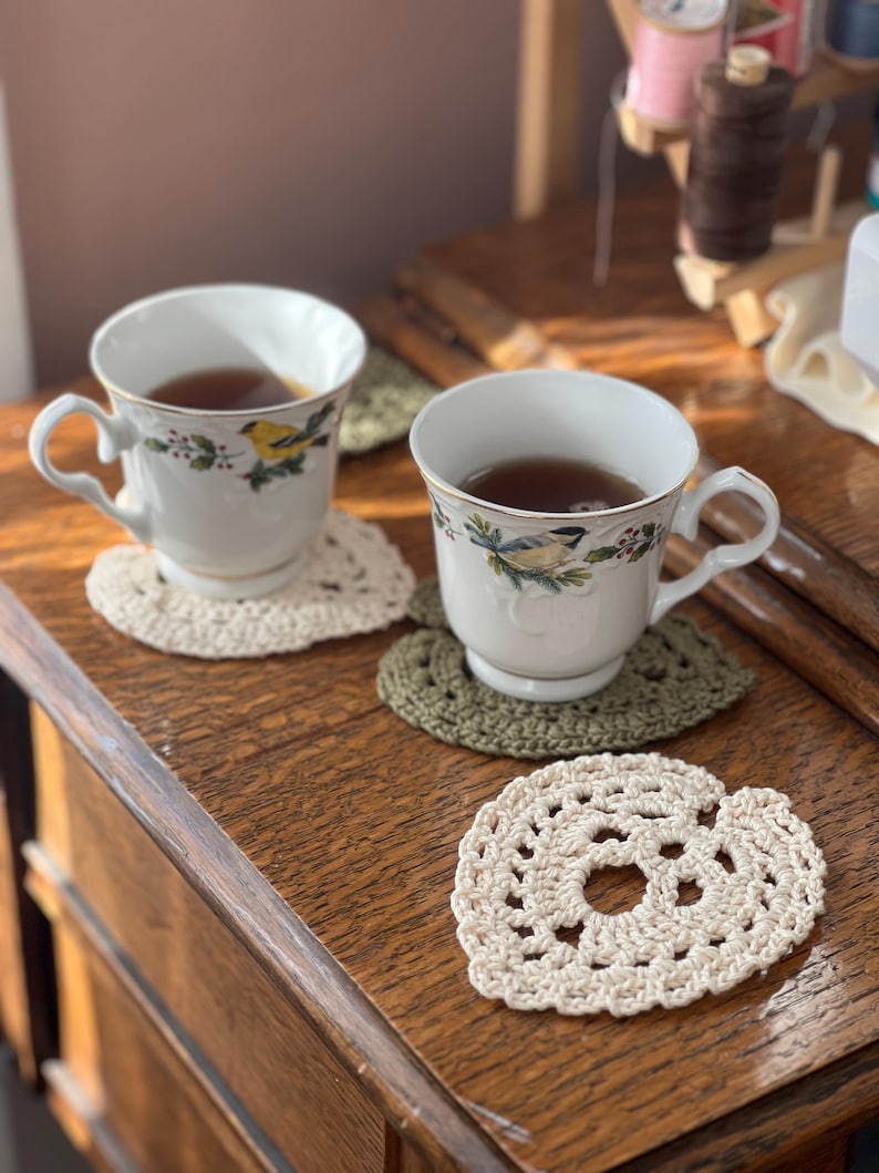 May include: Two white teacups with floral designs and brown tea inside, sitting on crocheted heart-shaped coasters on a wooden surface.