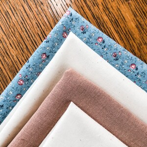 May include: Four fabric squares stacked on top of each other. The top square is a light blue fabric with a white floral pattern. The second square is a white fabric. The third square is a light brown fabric. The bottom square is a white fabric.