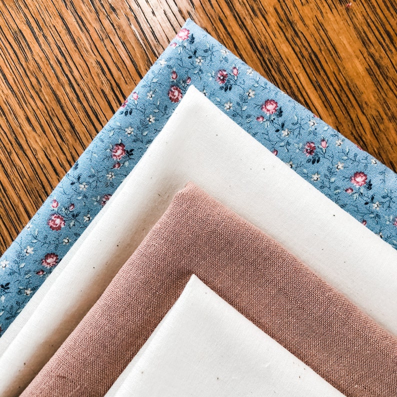 May include: Four fabric squares stacked on top of each other. The top square is a light blue fabric with a white floral pattern. The second square is a white fabric. The third square is a light brown fabric. The bottom square is a white fabric.