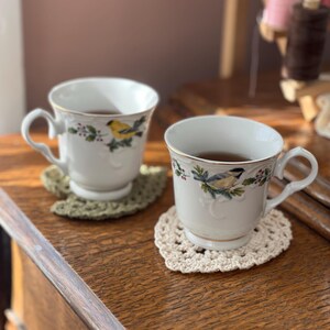 May include: Two white teacups with floral designs and gold trim, filled with tea, sitting on crocheted coasters. The coasters are in a heart shape and are beige and green.