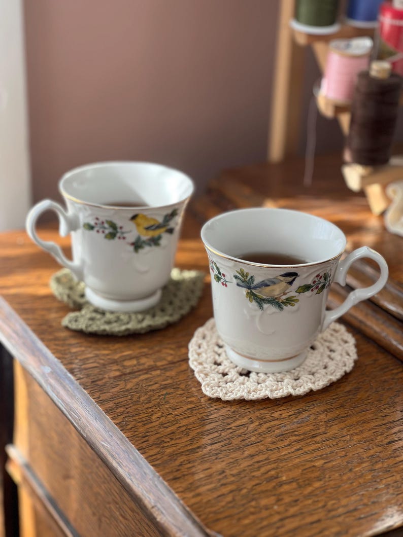 May include: Two white teacups with floral designs and gold trim, filled with tea, sitting on crocheted coasters. The coasters are in a heart shape and are beige and green.