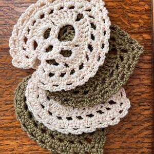 May include: A set of four crocheted coasters, two in a light beige color and two in a dark olive green color. The coasters are layered on top of each other, with the beige coasters on top.