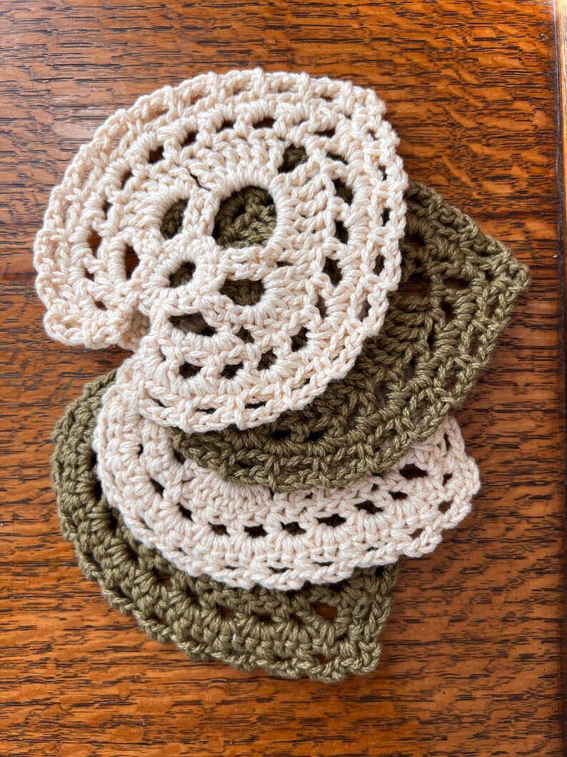 May include: A set of four crocheted coasters, two in a light beige color and two in a dark olive green color. The coasters are layered on top of each other, with the beige coasters on top.
