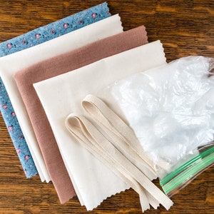 May include: A set of fabric scraps in various colors and patterns, including a blue floral print, a brown fabric, and a white fabric. There is also a bag of white stuffing and two pieces of white fabric ribbon.