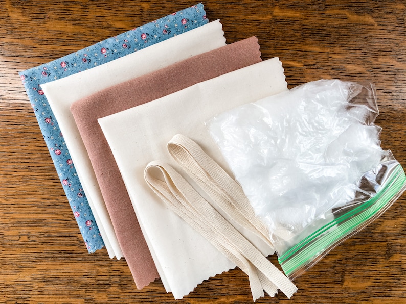 May include: A set of fabric scraps in various colors and patterns, including a blue floral print, a brown fabric, and a white fabric. There is also a bag of white stuffing and two pieces of white fabric ribbon.