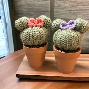 May include: Two crocheted cactus plants in terracotta pots with a pink and purple bow on each. The pots are sitting on a wooden tray.