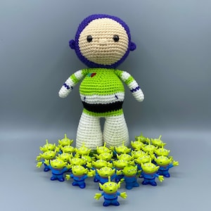 May include: A green and purple crocheted toy resembling a character from the Toy Story movie franchise. The toy is surrounded by a group of small green alien toys.