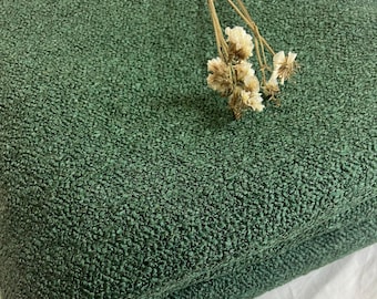 May include: A folded piece of textured, forest green fabric. The fabric has a bumpy, almost pebbled surface. A few sprigs of dried, cream-colored flowers rest on top of the fabric, which is lying on a white surface.