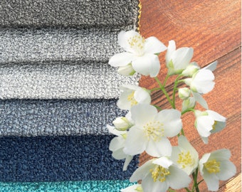 Op de afbeelding: Een close-up van vijf verschillende stofstalen in tinten grijs, blauw en turquoise. De stalen zijn gestapeld, met het grijze staal bovenop. Een boeket witte jasmijn bloemen is rechts in de afbeelding geplaatst, gedeeltelijk de stofstalen bedekken.
