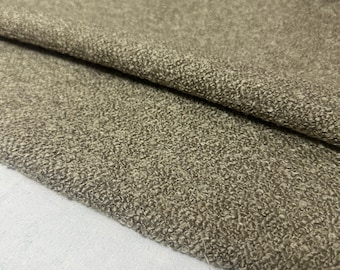 Op de afbeelding: Close-up van een getextureerde stof in tinten bruin en beige. De stof heeft een noppen, geluste textuur, met plooien en schaduwen die diepte toevoegen. Het materiaal lijkt zacht en is mogelijk geschikt voor bekleding of kleding.