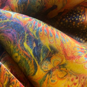 Op de afbeelding: Een close-up van een kleurrijke stof met een paisley-patroon. De stof is een mix van geel, oranje, rood, blauw en groen. Het patroon bevat pauwen, vlinders en bloemenmotieven.