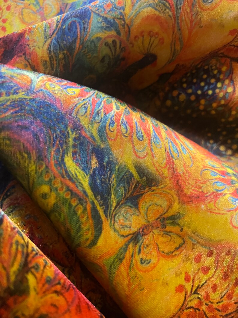 Op de afbeelding: Een close-up van een kleurrijke stof met een paisley-patroon. De stof is een mix van geel, oranje, rood, blauw en groen. Het patroon bevat pauwen, vlinders en bloemenmotieven.