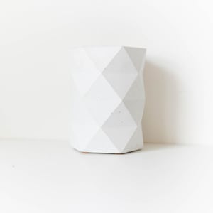 May include: A white, geometric, cylindrical planter with a diamond pattern. The planter is made of a textured material and has a small, light brown base. The design is modern and minimalist, suitable for home decor.