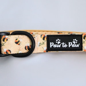 Peut inclure: Un collier de chien jaune avec une boucle noire et un motif d'abeilles noires et blanches. Le collier a une étiquette noire qui dit "Paw to Paw".
