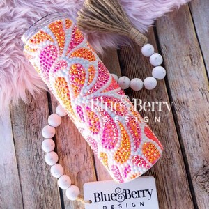 May include: A cylindrical tumbler covered in orange, pink, and white rhinestones arranged in a swirling paisley pattern. The tumbler features a clear lid and is accompanied by a string of white wooden beads with a jute tassel and a white tag labeled Blue Berry DESIGN.