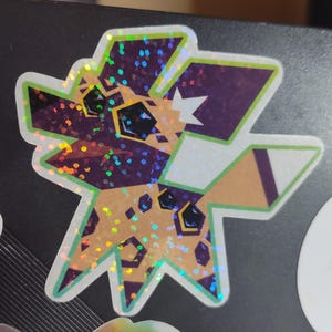 May include: A holographic sticker featuring a geometric design of a stylized animal head. The sticker has a purple, tan, and white color scheme with green and iridescent accents. The animal's eyes are black.