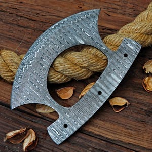 Op de afbeelding: Een Damascus stalen bijlkop met een unieke halve maanvorm en een patroon. De bijlkop heeft een grote cirkelvormige opening en drie gaten. Het ligt op een houten oppervlak met touw en gedroogde zaaddozen.