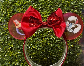 May include: A headband featuring two circular ears and a large red satin bow with a glittery center. The left ear displays a cartoon character with long red hair and purple gloves, while the right ear features a white cartoon rabbit character.