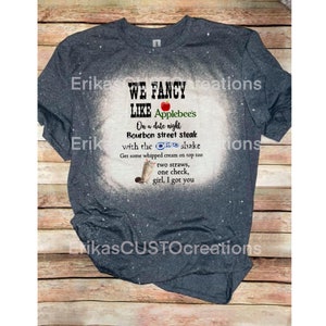 Puede incluir: Camiseta azul marino con un diseño blanco decolorado. La camiseta presenta el texto "WE FANCY LIKE Applebee's" junto con una lista de elementos del menú y un gráfico de batido. Es una prenda informal.