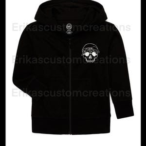 Puede incluir: Sudadera negra con cremallera y un gráfico de calavera blanca con pañuelo. La sudadera con capucha tiene una capucha y una cremallera de largo completo. El gráfico de la calavera está en el área del pecho izquierdo. La marca es "Wonder Nation".