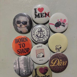 May include: A collection of round pinback buttons with various designs. Some buttons feature text like "I ❤️ MEN", "BORN TO SHOP", and "Diva". Other buttons display images of a dog wearing sunglasses, Snoopy, and a sailing club logo.