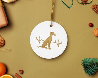Op de afbeelding: Een wit keramiek ornament met een gouden ophangkoord. Het heeft een bruinachtige hondenillustratie en een hartslaglijn ontwerp. De achtergrond is een gedempt groen met decoraties.
