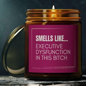 Peut inclure: Une bougie en pot de verre brun avec un couvercle doré et une mèche allumée. L'étiquette est magenta avec un texte blanc qui dit "SMELLS LIKE... EXECUTIVE DYSFUNCTION IN THIS BITCH." La bougie est coulée à la main avec de la cire de noix de coco.