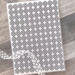 Puede incluir: Una carpeta de relieve rectangular de plástico transparente con un patrón de trébol negro repetido. El diseño es una serie de formas de tréboles de cuatro hojas entrelazadas. Una cinta blanca con pequeños lunares es visible en la esquina inferior izquierda.